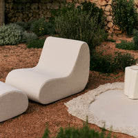 "White, curved patio chair and ottoman on a natural, textured ground with green foliage in the background."