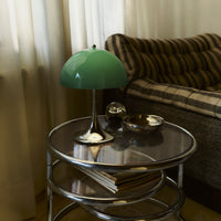 Green lamp atop a silver table, accompanied by dark bowls, with a striped brown and beige couch in the background.