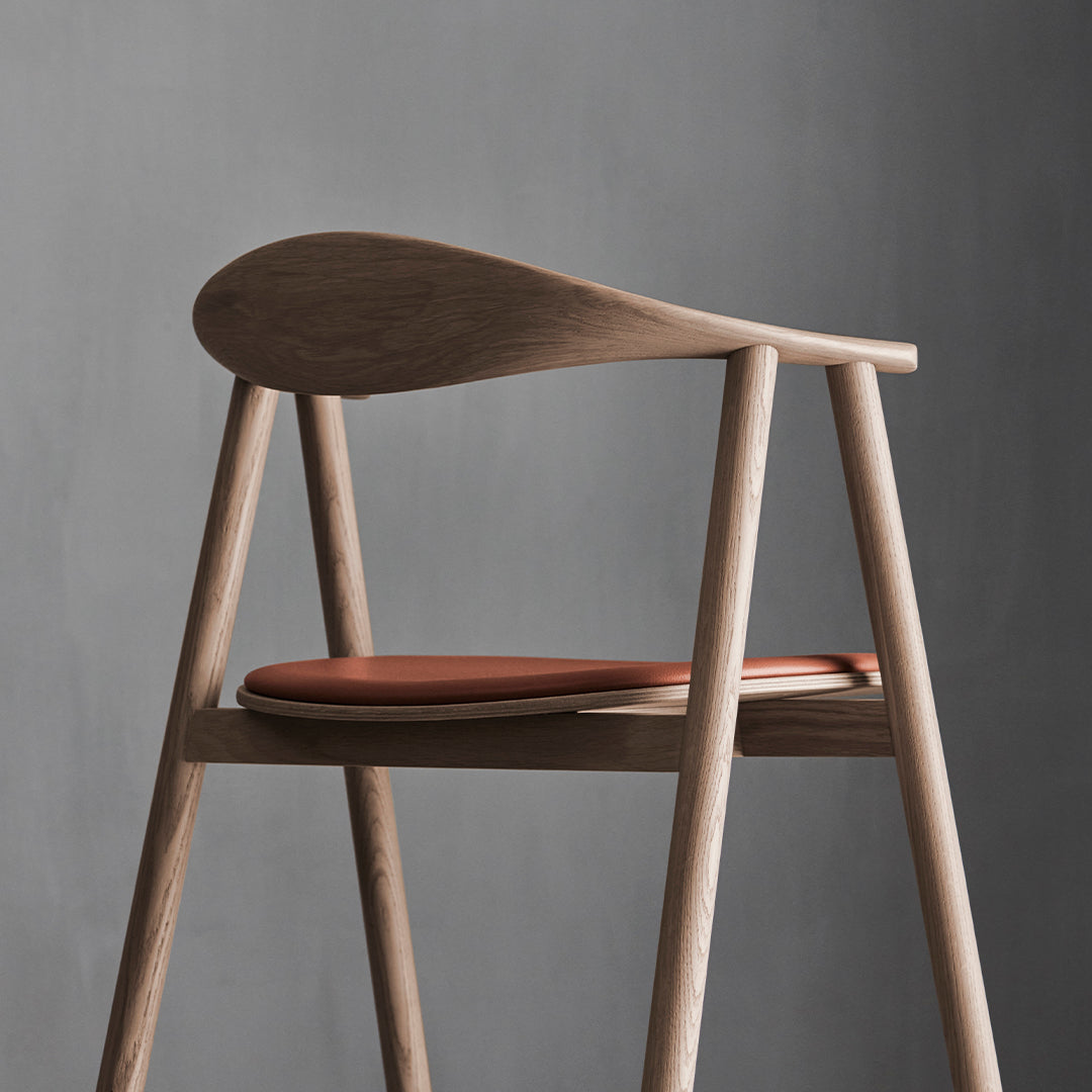 Minimalist wooden chair with a light ash finish and a deep brown leather seat against a gray background.