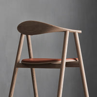 Minimalist wooden chair with a light ash finish and a deep brown leather seat against a gray background.