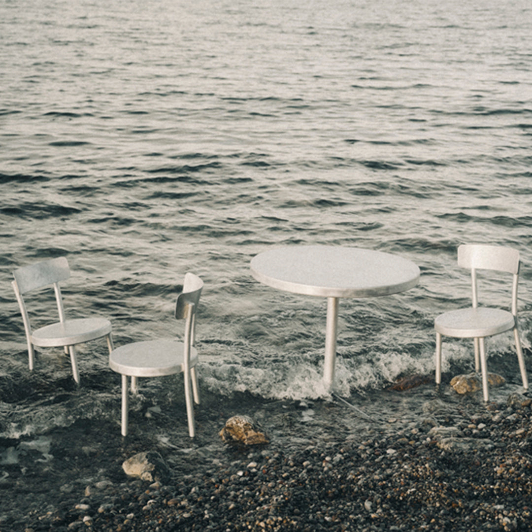 Silver metal outdoor dining set with a round table and four matching chairs near a serene waterline.