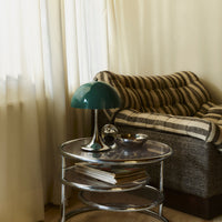 Green lamp on a chrome table beside a striped brown and beige sofa, with light curtains in a cozy interior setting.