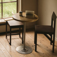 Round wooden table with a light finish, paired with two dark wooden chairs on a natural wood floor.