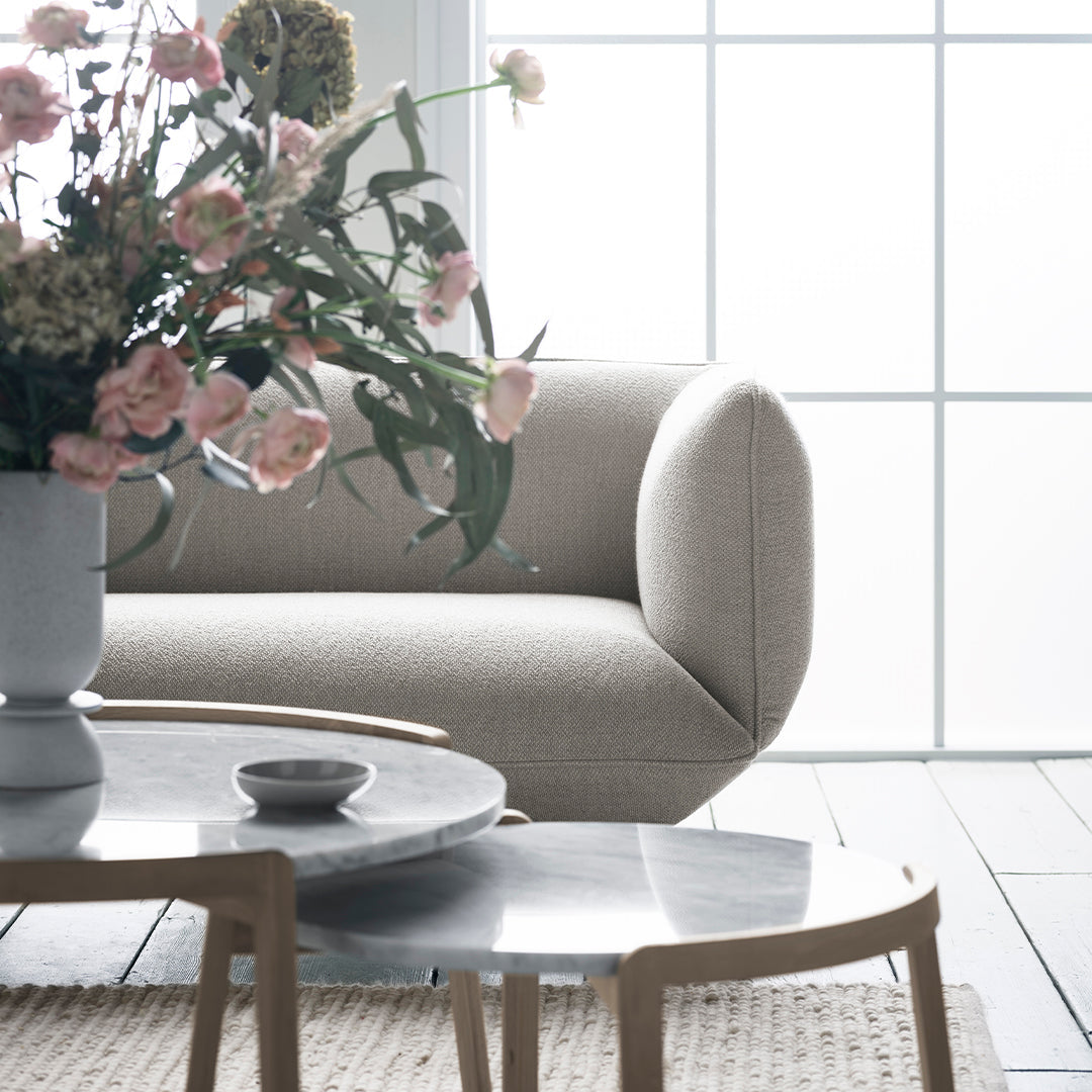 Soft beige sofa with a smooth texture, complemented by a gray marble table and pastel floral arrangement. Bright, airy ambiance.