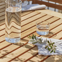 Clear glassware on a light wooden table with natural wood grain, featuring olive green leaves and white napkins.