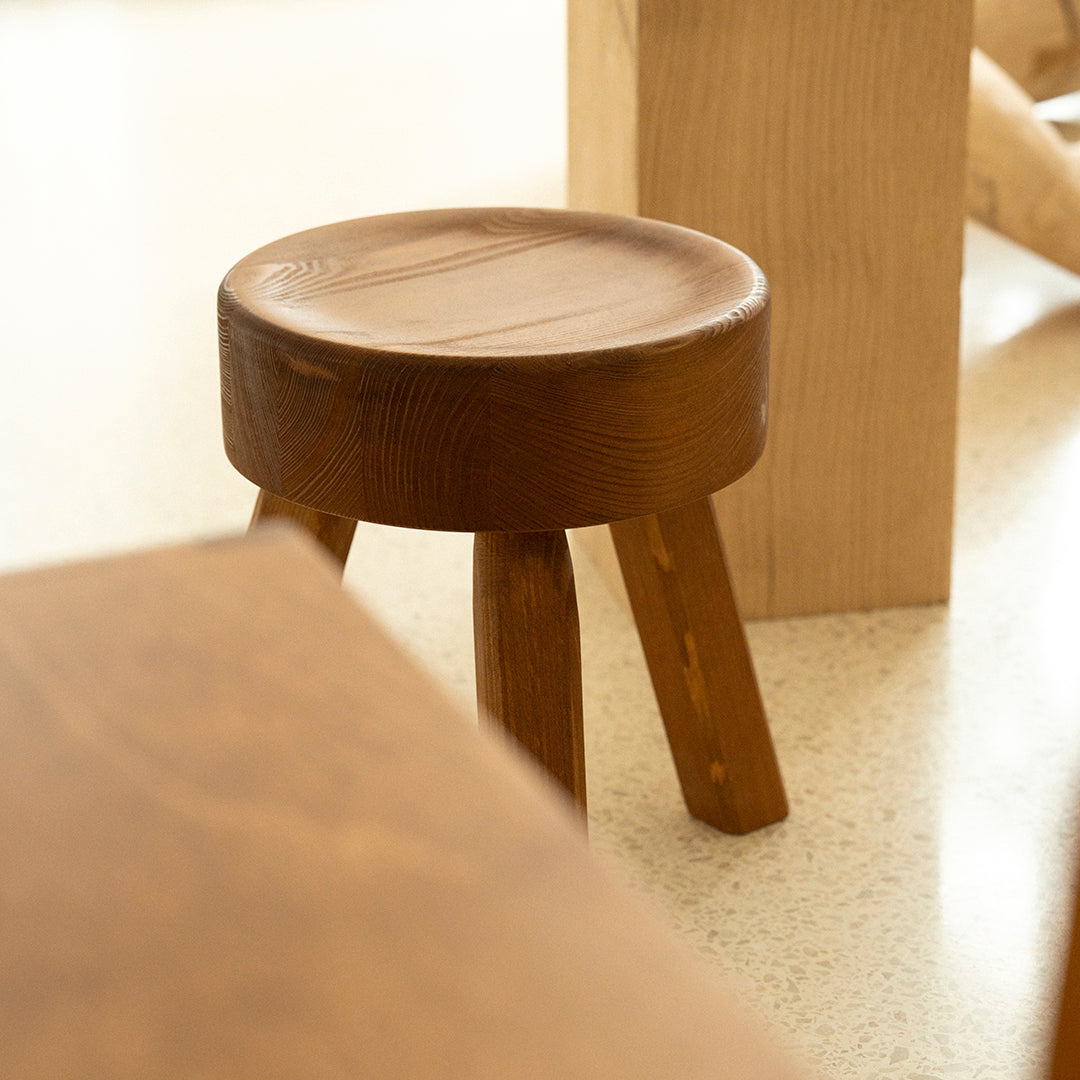 Brown wooden stool with a smooth, circular seat and three tapered legs, showcasing natural wood grain patterns.