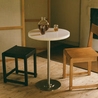 Minimalist white round table with a stainless steel base, surrounded by a black stool and a natural wood chair.
