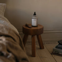 Round wooden side table with natural wood finish and three tapered legs, set on a light wooden floor. Simple and modern design.