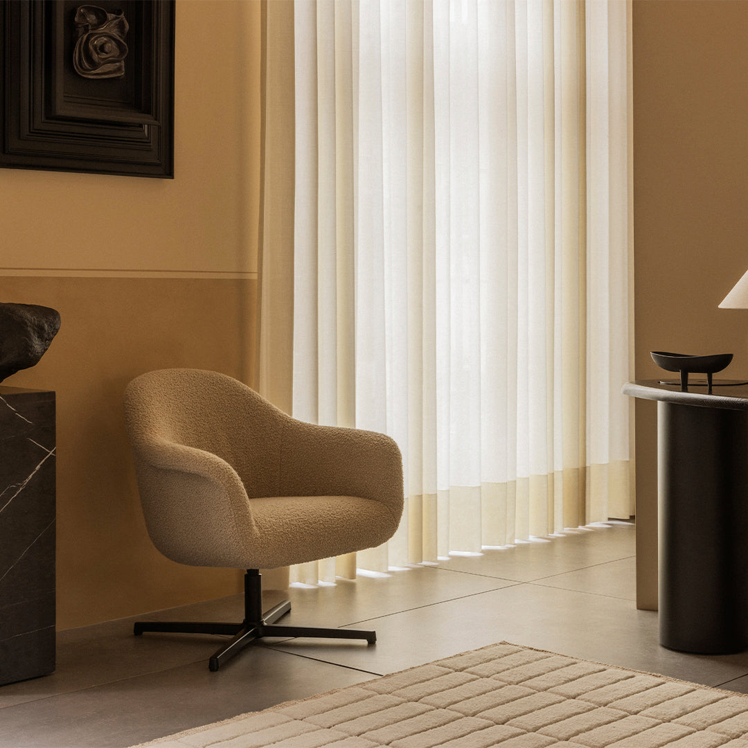 Beige curved chair with a soft texture, set against light cream curtains and a neutral-toned patterned rug.