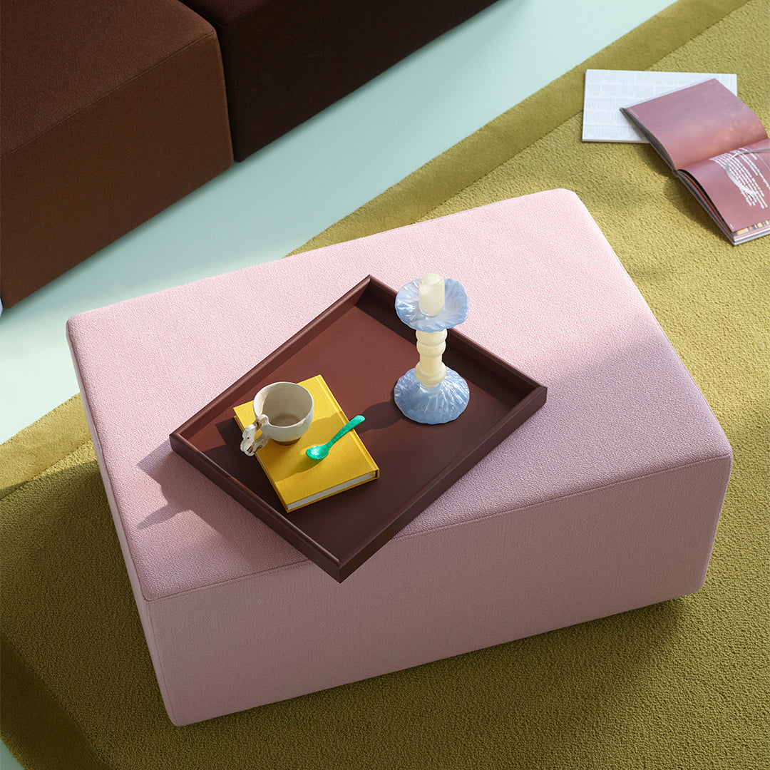 Pink ottoman atop a green rug, styled with a brown tray, a yellow book, a blue candle holder, and a ceramic cup.