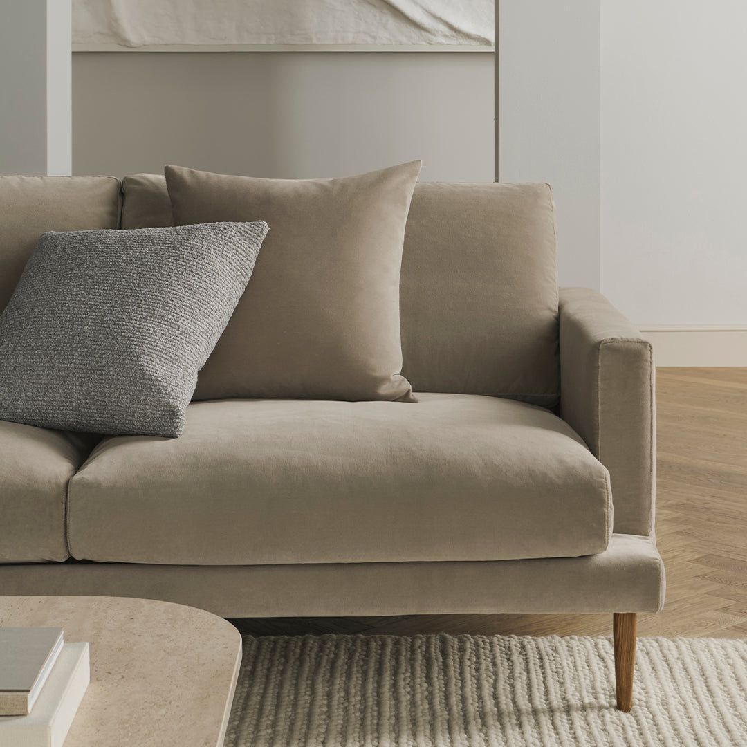 Beige couch with a soft texture, featuring a dark gray accent pillow and light wooden legs, set against a neutral backdrop.