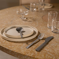 Neutral beige plates on a textured, golden table with sleek silver utensils and clear glassware. Elegant, modern design elements.