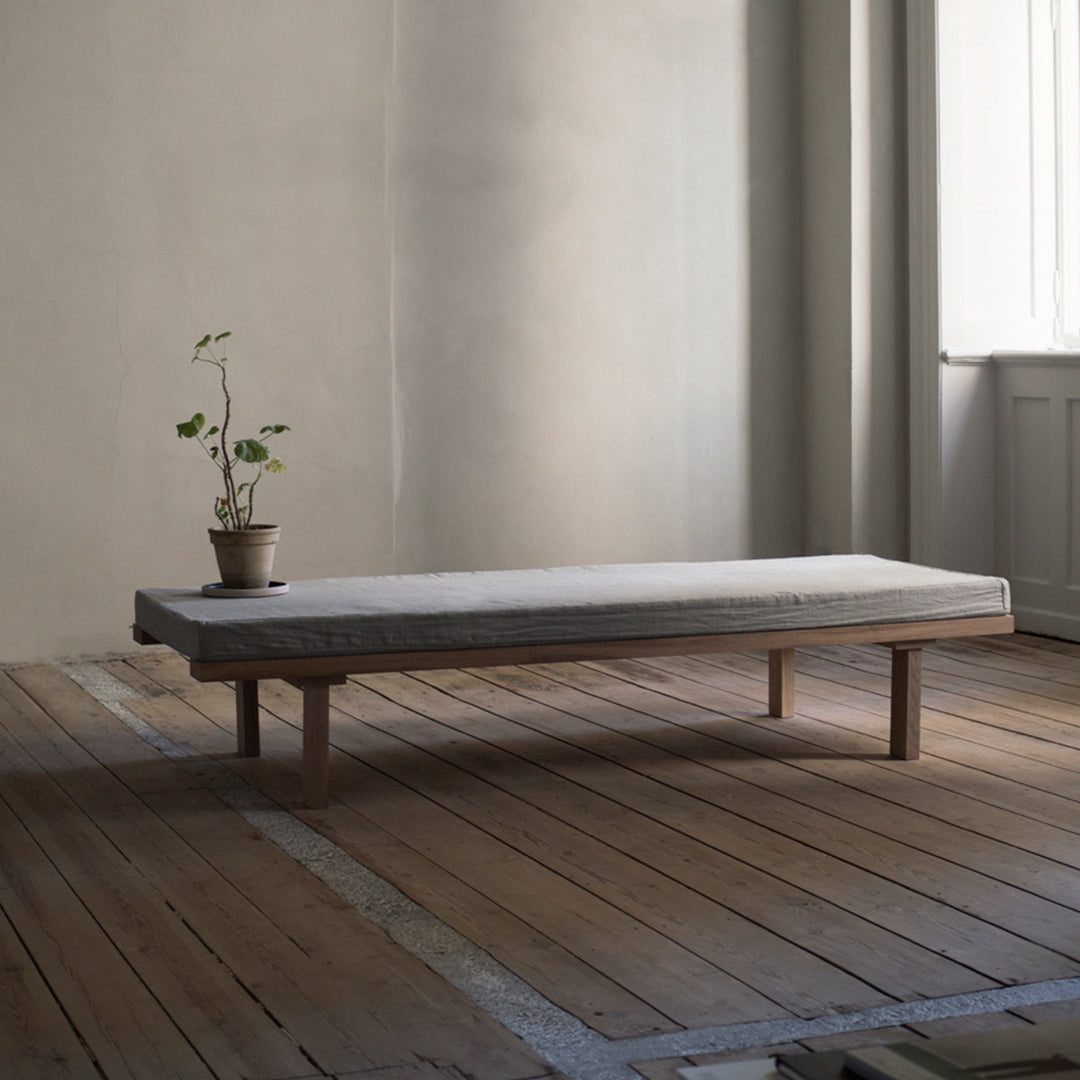 Minimalist gray mattress on a natural wood platform, surrounded by a neutral-toned interior and light wood flooring.