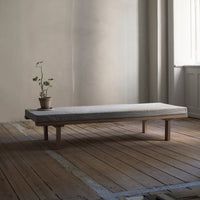 Minimalist gray mattress on a natural wood platform, surrounded by a neutral-toned interior and light wood flooring.