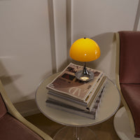 Yellow lamp on a glass table with stacked magazines, surrounded by pink armchairs in a modern, stylish setting.
