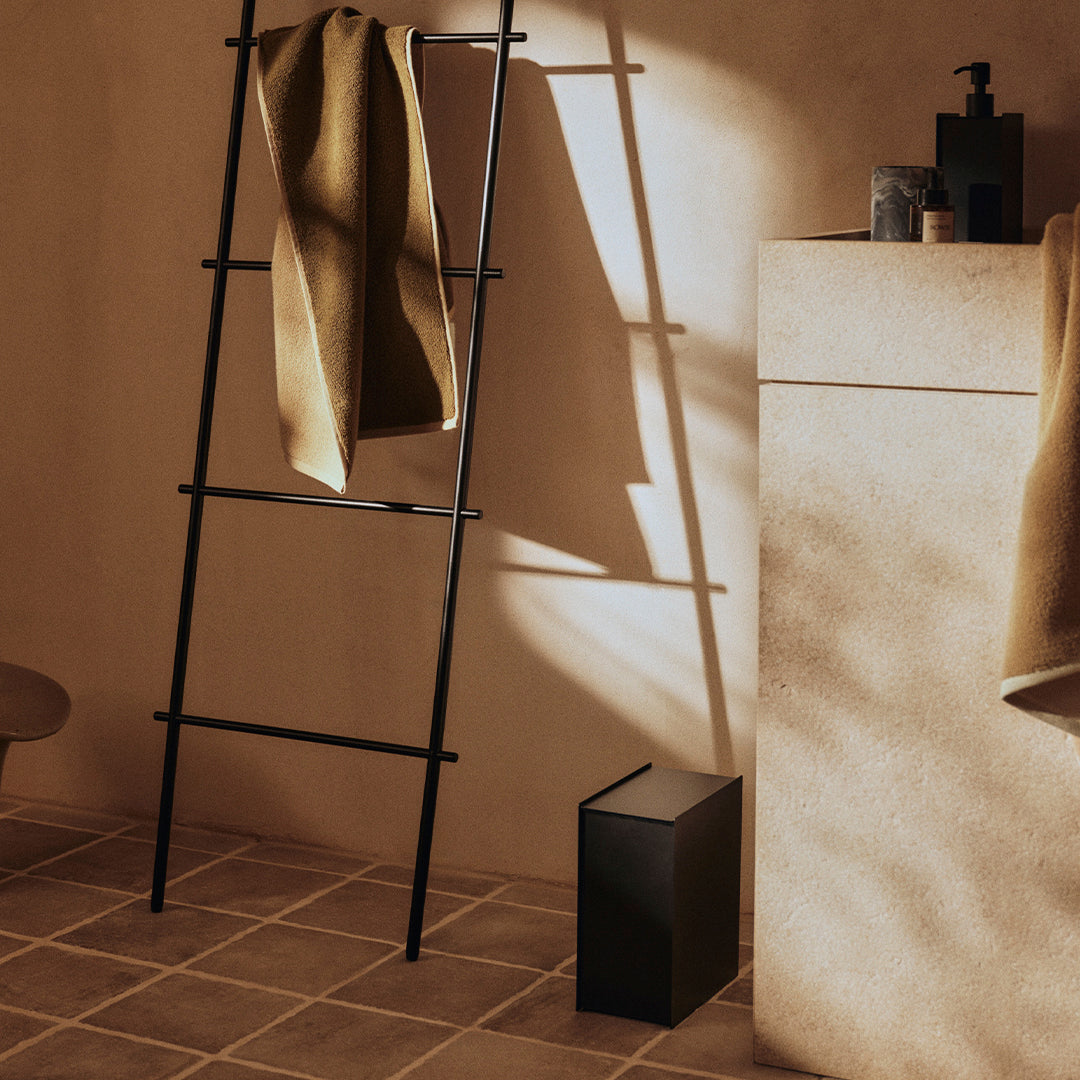 "Minimalist bathroom scene with earthy tones, black ladder, and beige textured walls casting soft shadows."