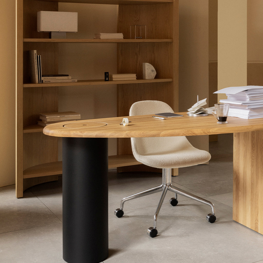 Natural wood desk with a light beige chair and black cylindrical base, set against a warm-toned interior.