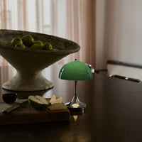 Green glass lamp with a shiny chrome base, set against a neutral backdrop featuring a textured bowl of green apples.