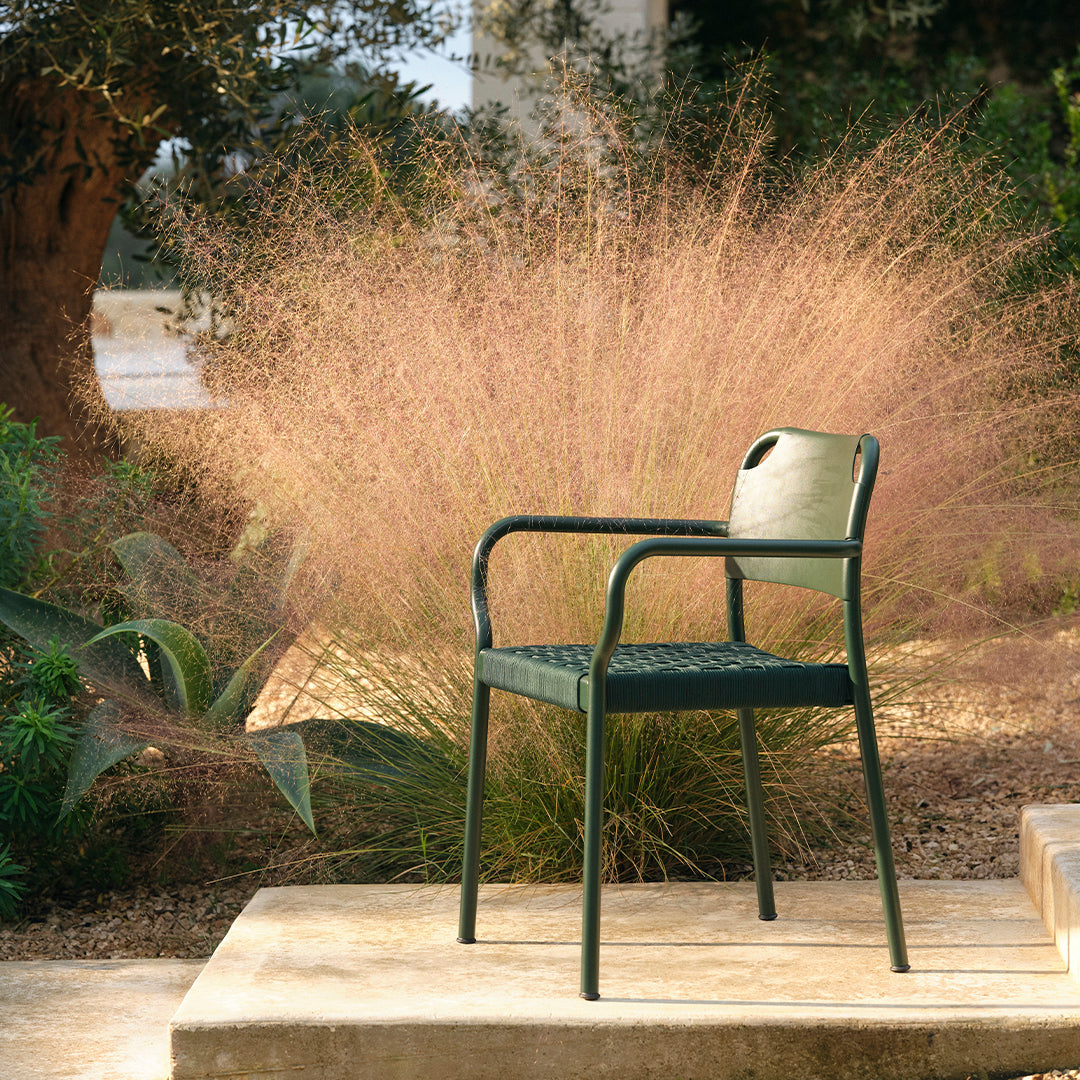 Dark green armchair with a textured pattern, set against a backdrop of soft pink grasses and lush greenery.