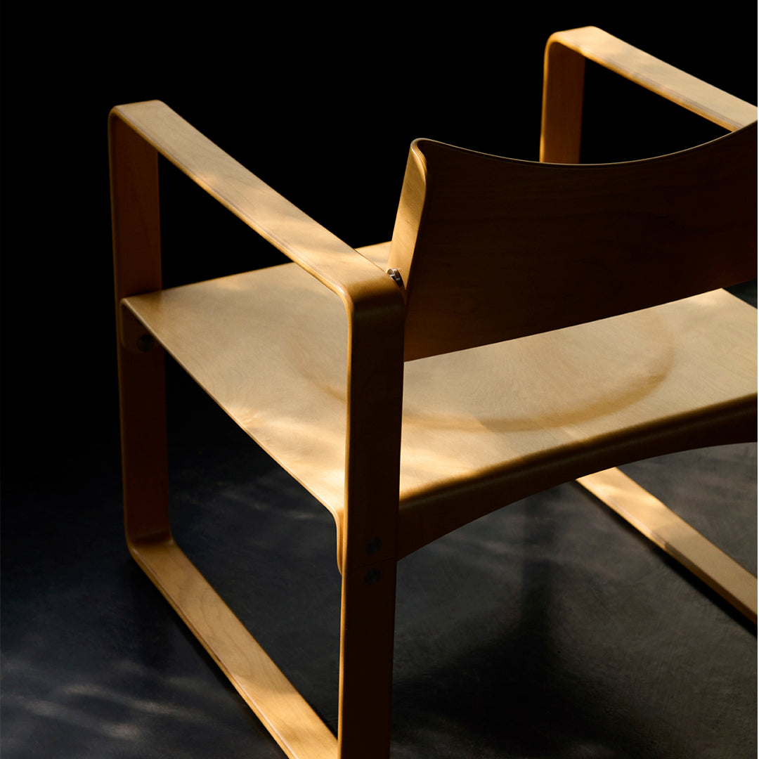 Light wood chair with minimalistic design; smooth, natural finish; shadows create a subtle pattern against a dark background.