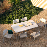 Rectangular outdoor dining table with light gray surface and patterned gray chairs, set in a lush garden with greenery.