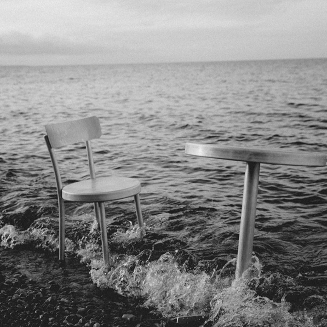 Wooden chair and round table by the water, set against a serene black and white backdrop with gentle waves.