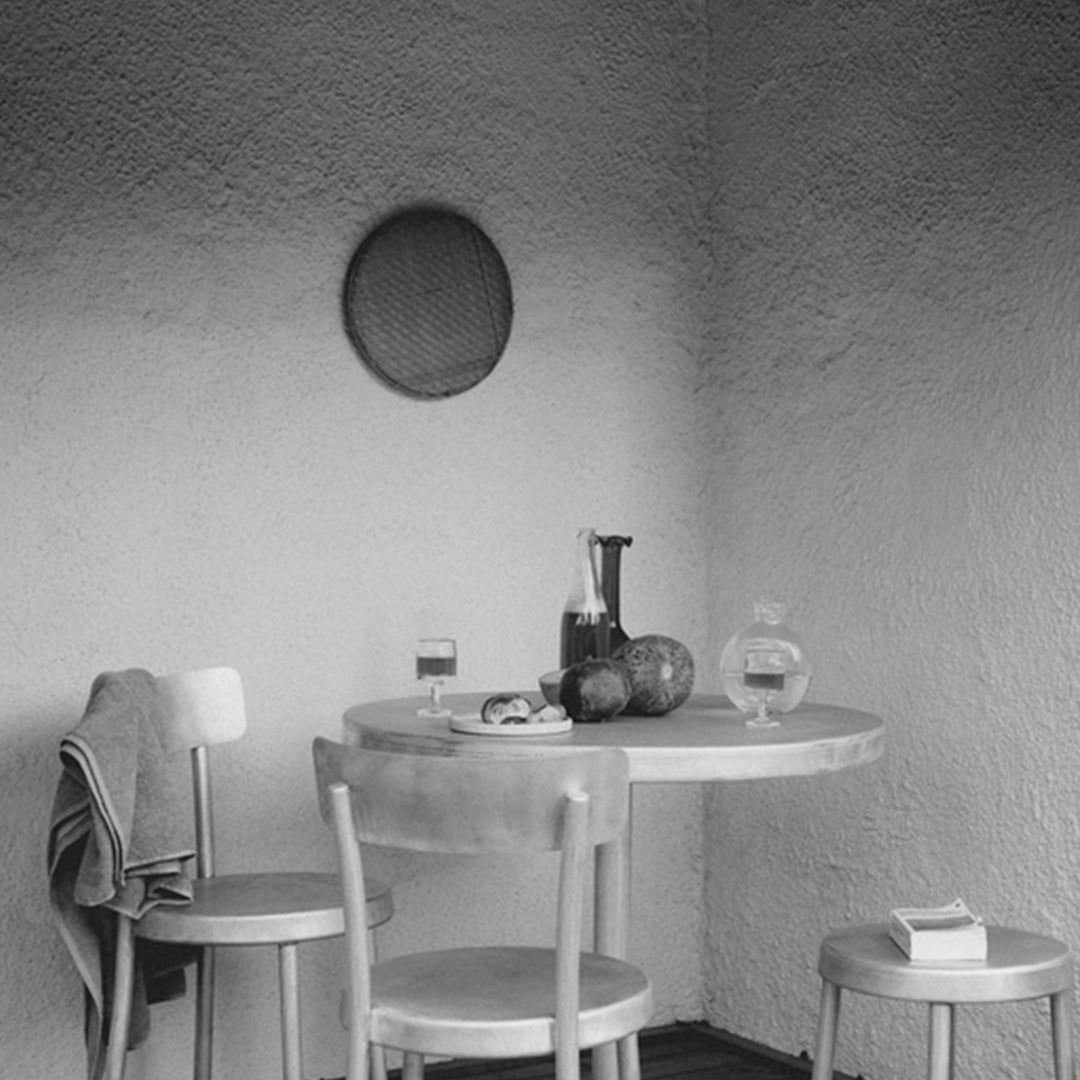 Black and white scene of a minimalist table setting with wooden chairs and simple decor on a textured wall.