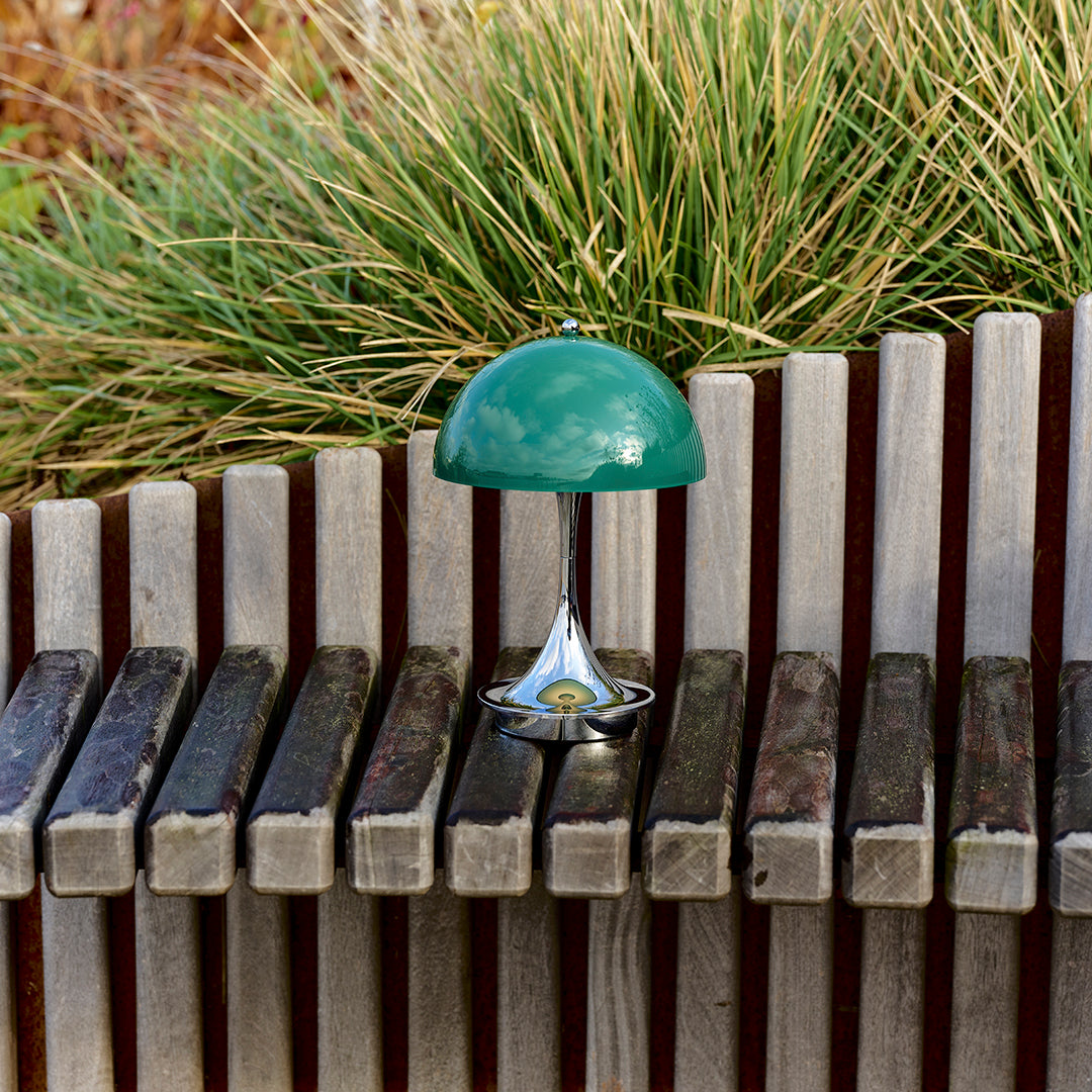 Green dome lamp with a sleek chrome base, set against wooden slats and lush greenery. Modern design and vibrant colors.