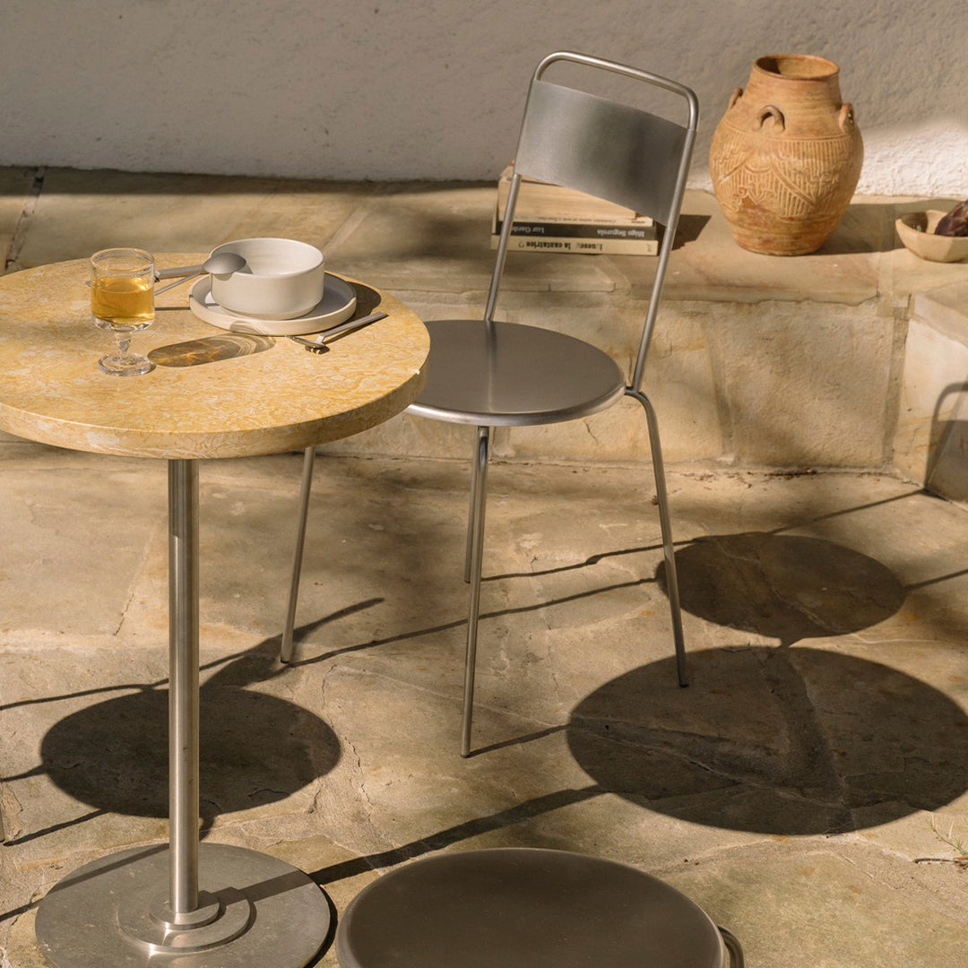 "Round stone-patterned table with silver chair and shadows, set against a natural stone floor and clay pottery."