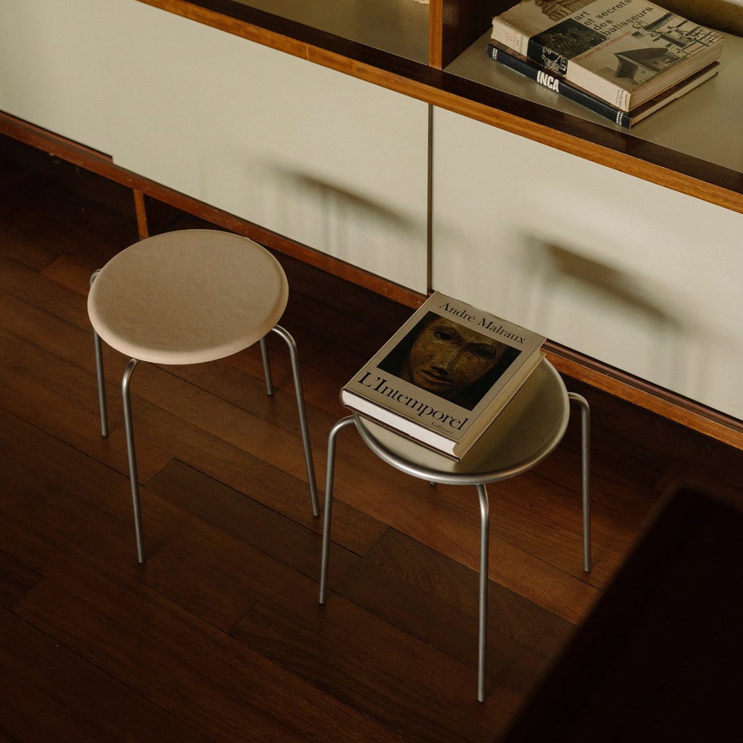 Two minimalist stools with round beige tops and sleek silver legs, placed on warm wooden flooring.