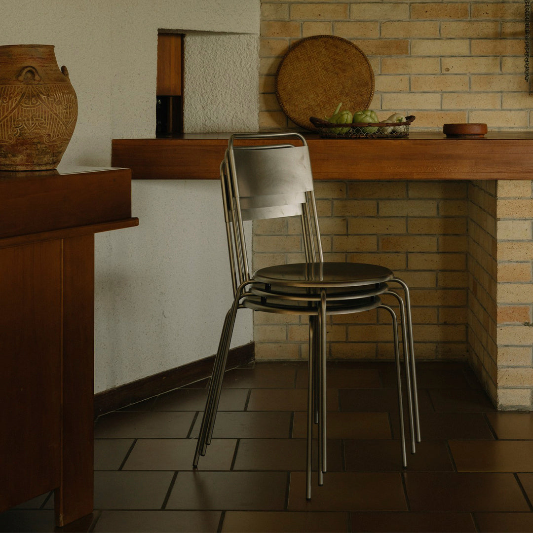 Stackable chair in sleek metallic finish, featuring simple lines and subtle reflections, set against warm wood and brick tones.