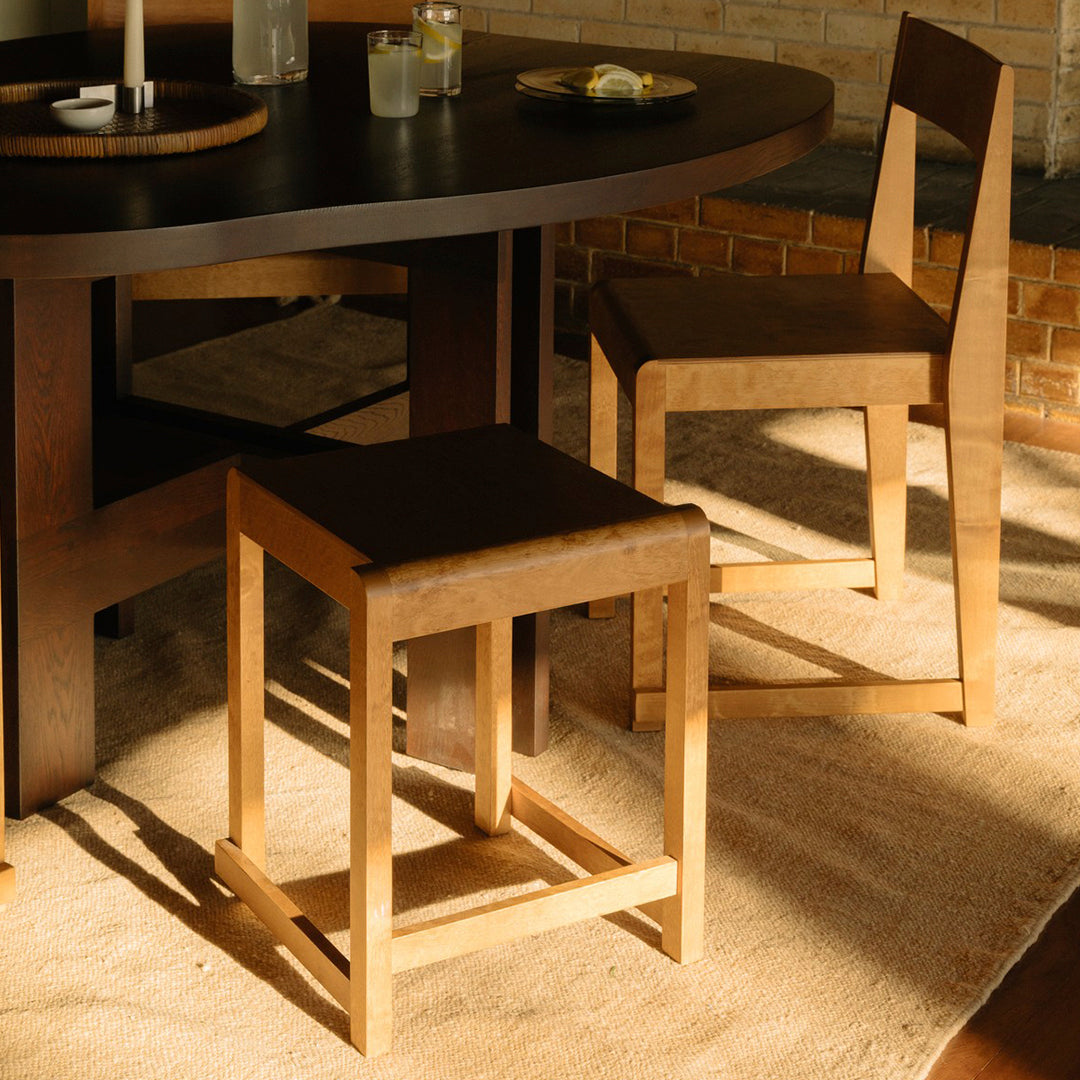 Natural wood stool in light and dark brown tones, set against a warm beige rug and dark round table.