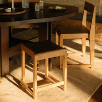 Natural wood stool in light and dark brown tones, set against a warm beige rug and dark round table.