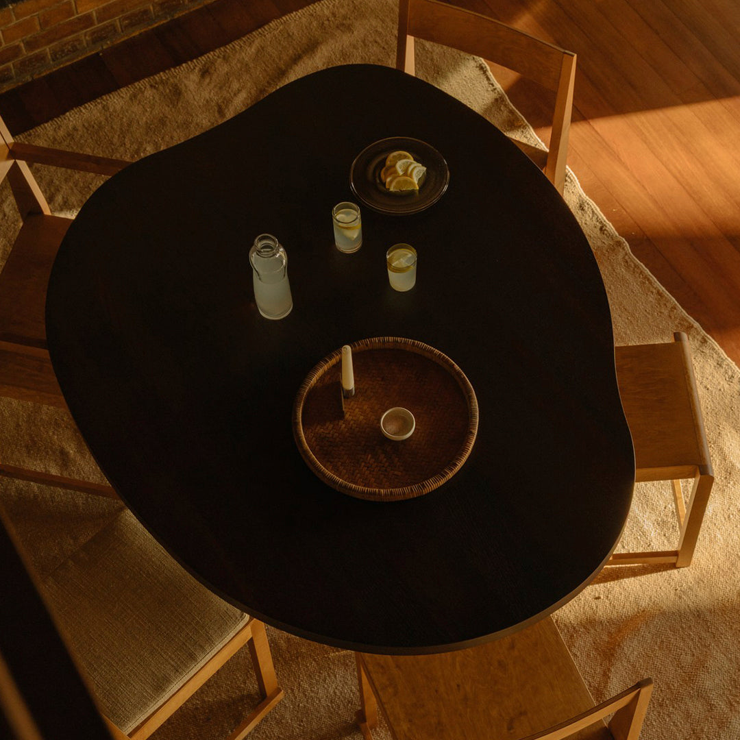 "Dark organic-shaped table with wooden bowl and round plates, surrounded by light-toned chairs and soft textured rug."