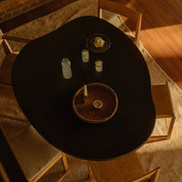 "Dark organic-shaped table with wooden bowl and round plates, surrounded by light-toned chairs and soft textured rug."