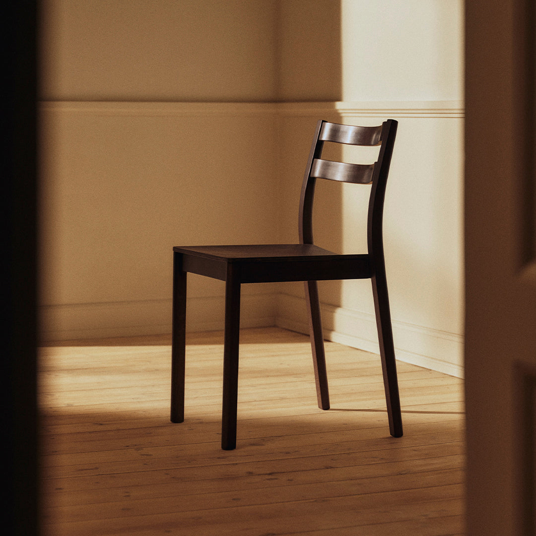 "Elegant black wooden chair with a simple slatted backrest, set against a warm-toned, sunlit interior."