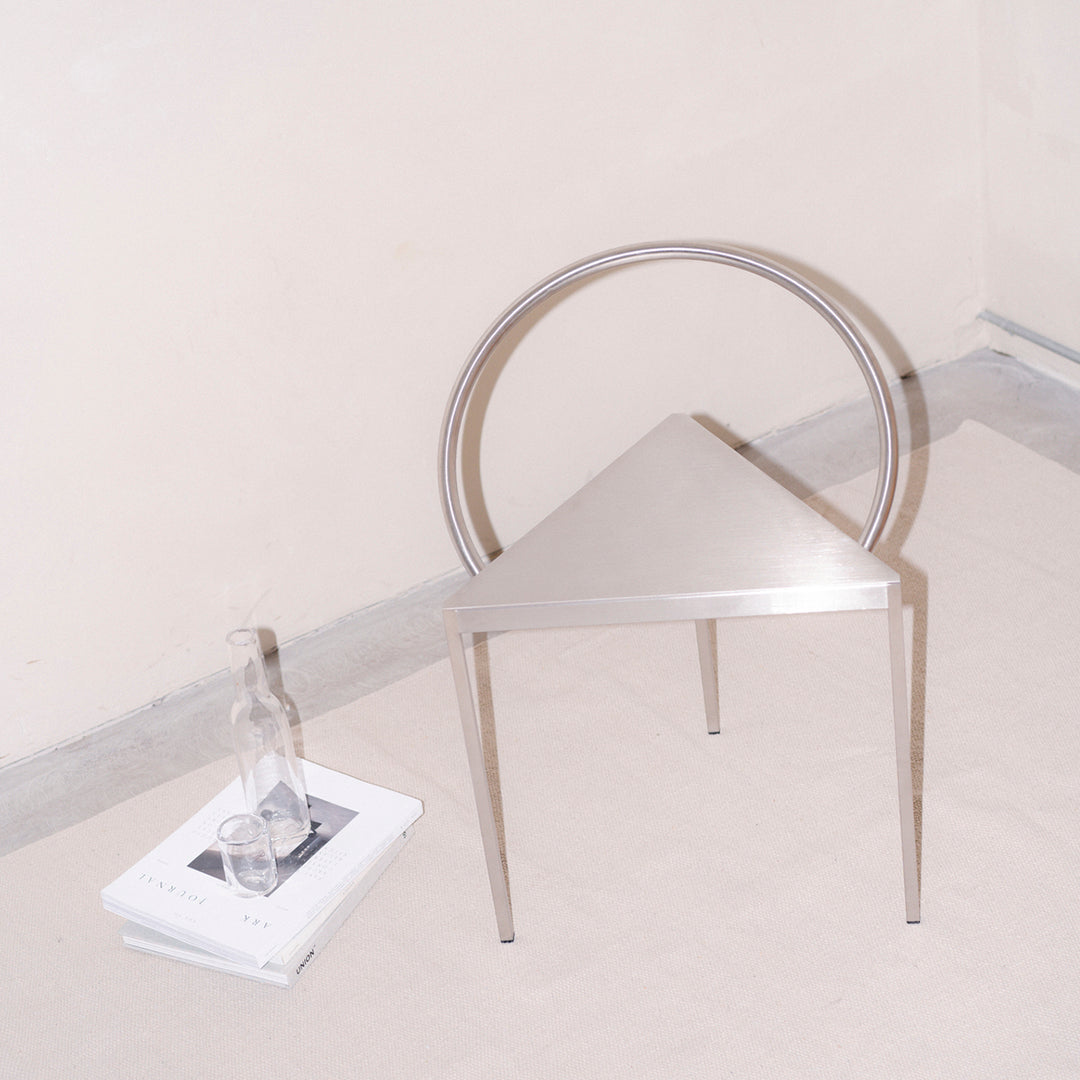 Triangular silver chair with circular back, minimalist design, and sleek metallic finish against a neutral background.
