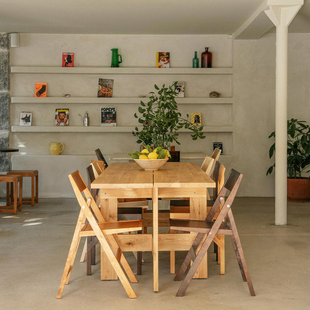 Wooden dining table with light and dark brown folding chairs, complemented by greenery and neutral-colored walls.