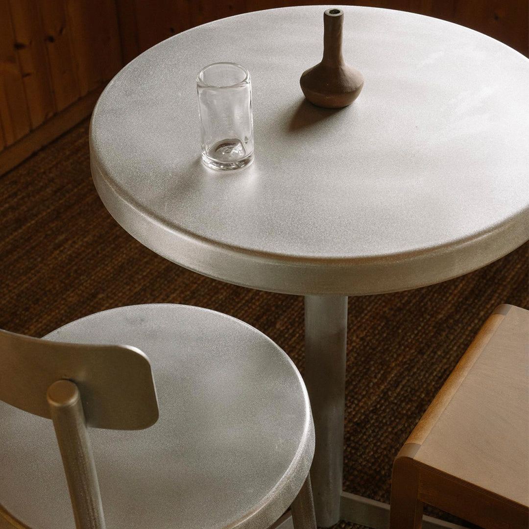 Round silver table with smooth finish, paired with a light wooden chair, creating a minimalist and modern aesthetic.