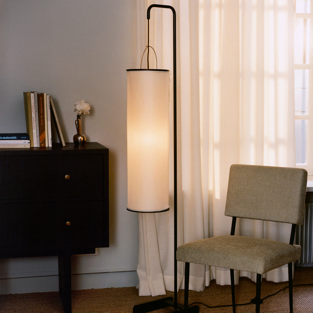 Modern black and white floor lamp with a cylindrical shade, set against a neutral gray wall and light curtains.