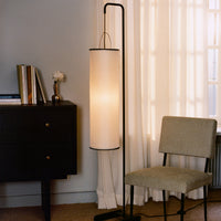 Modern black and white floor lamp with a cylindrical shade, set against a neutral gray wall and light curtains.
