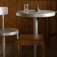 Elegant round metallic table with smooth silver finish, paired with a warm wood square stool. Rustic wooden backdrop.