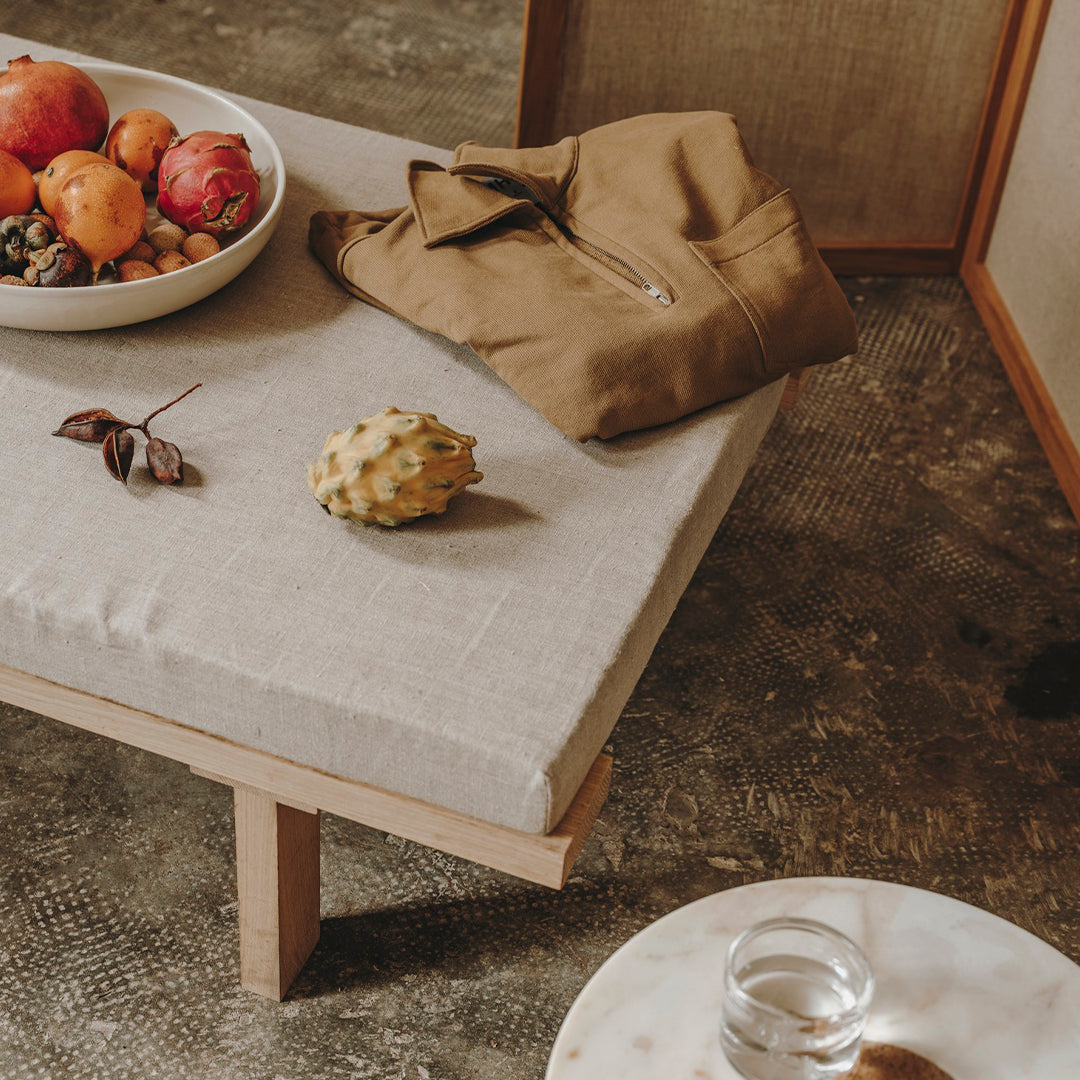 Beige cushioned bench with wooden legs, surrounded by a fruit bowl, brown jacket, and a marble coffee table.