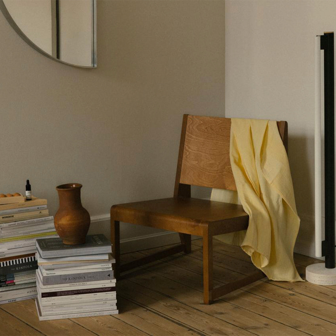 Wooden chair with a yellow draped fabric, stacked white books, and a brown vase in a neutral-toned room.