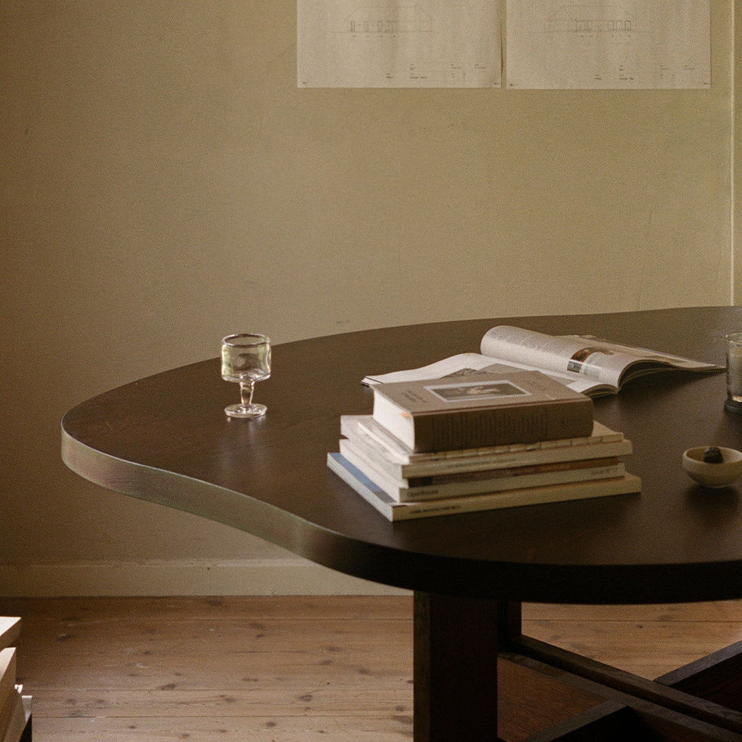 Curved dark wood table with stacked books and a glass, featuring warm neutral tones and minimalist design.