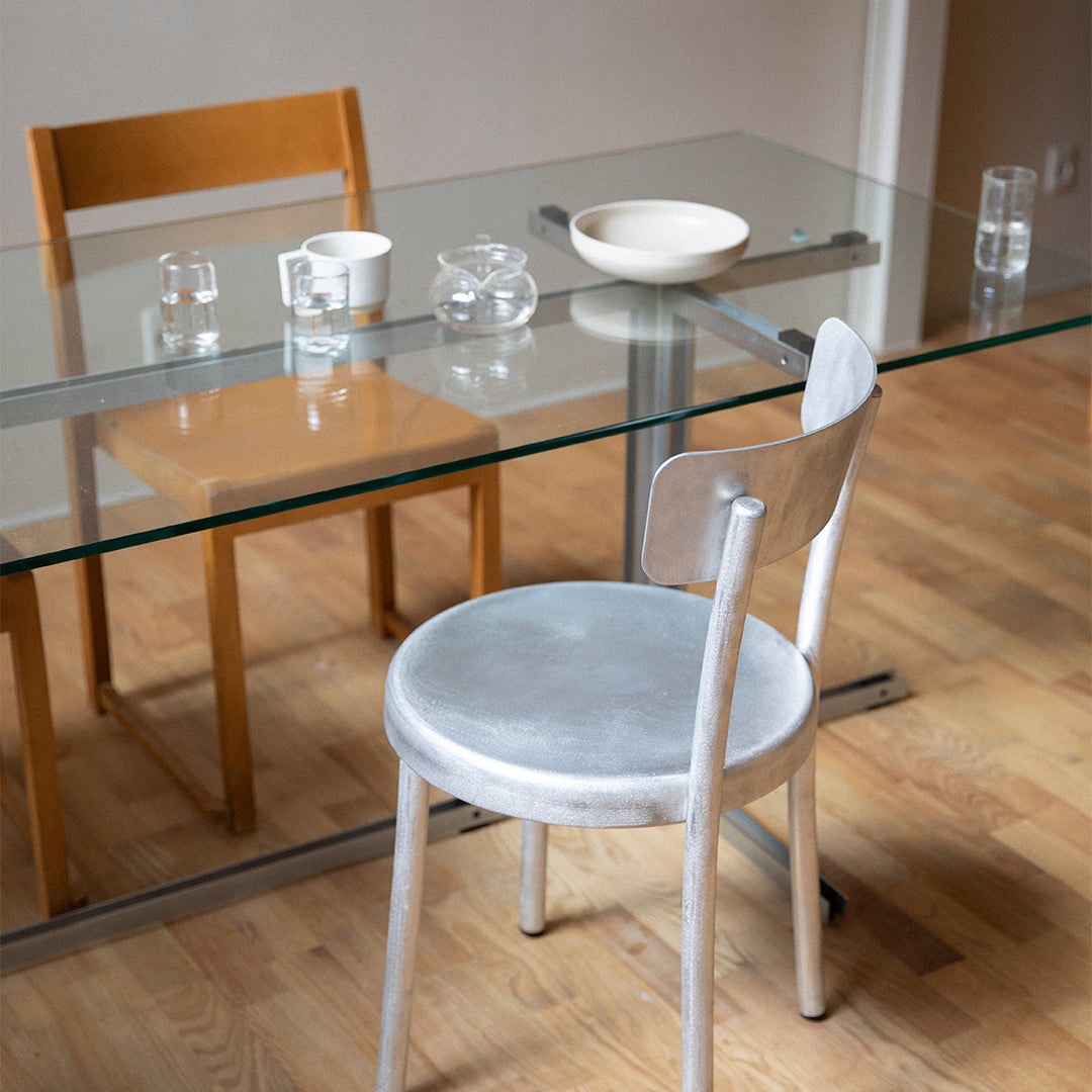 Gray velvet chair beside a glass table, highlighted by wooden chair and minimalist dishware on a light wood floor.