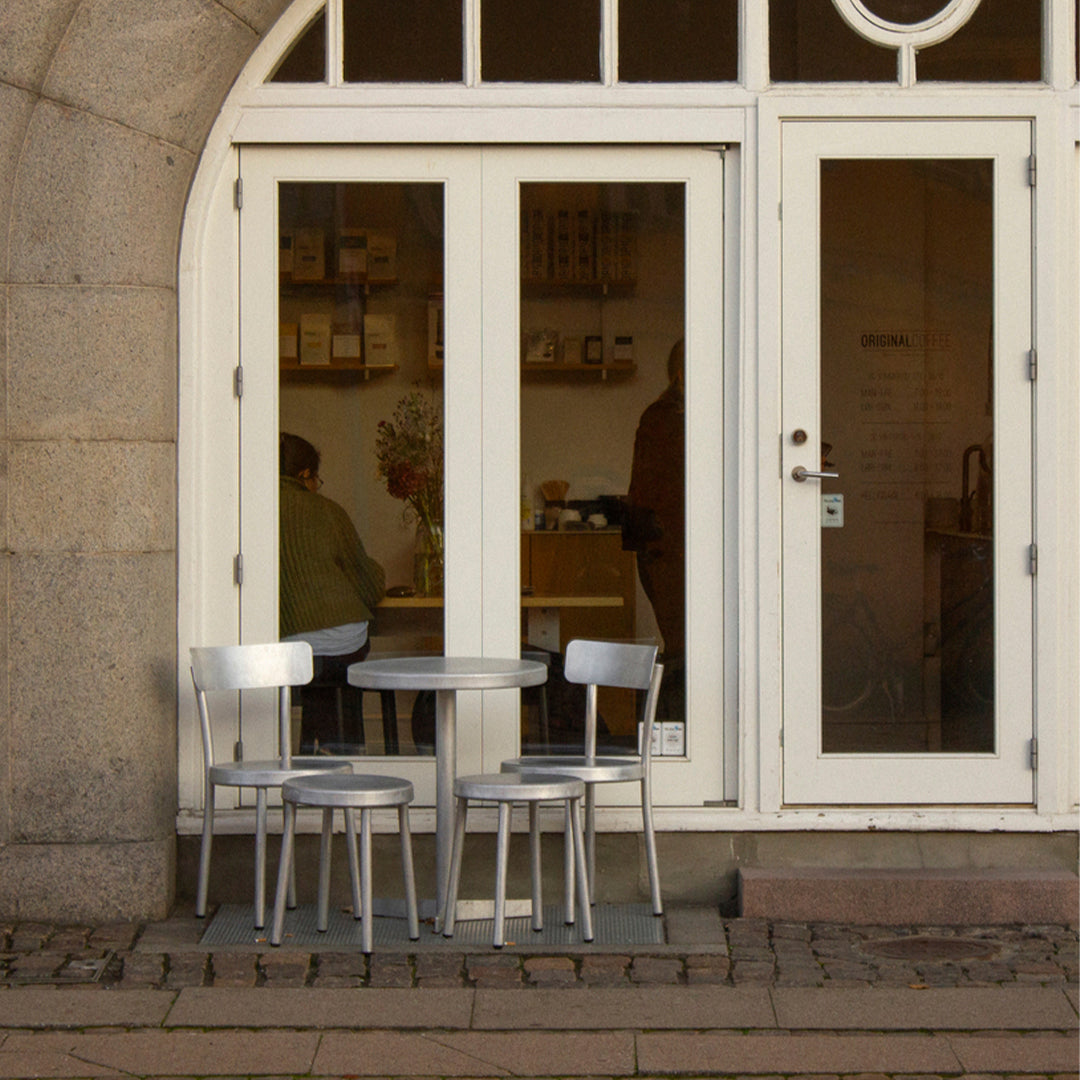 Silver metal chairs and table set against a light cream-colored entrance, with a hint of rustic stone texture.