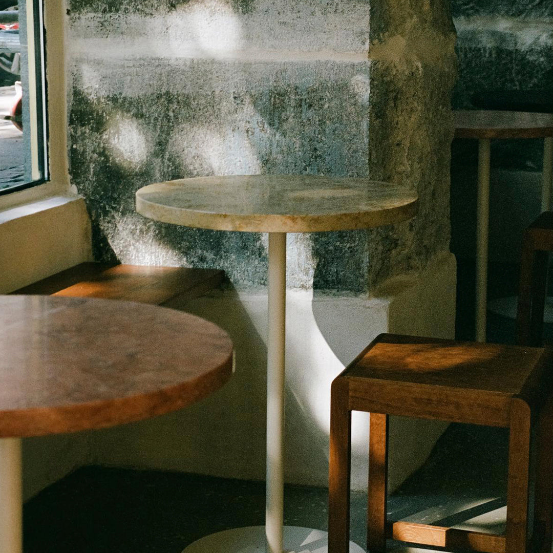 Light-colored round tabletop with subtle marble pattern, paired with dark wooden square stools, against a textured gray wall.