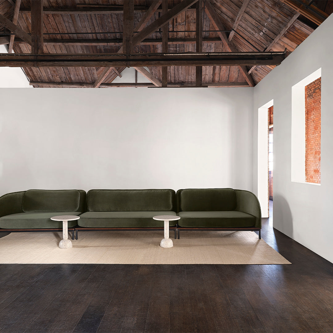 Sleek dark green velvet sofa with a light-colored round side table, complemented by a neutral rug on dark wood flooring.
