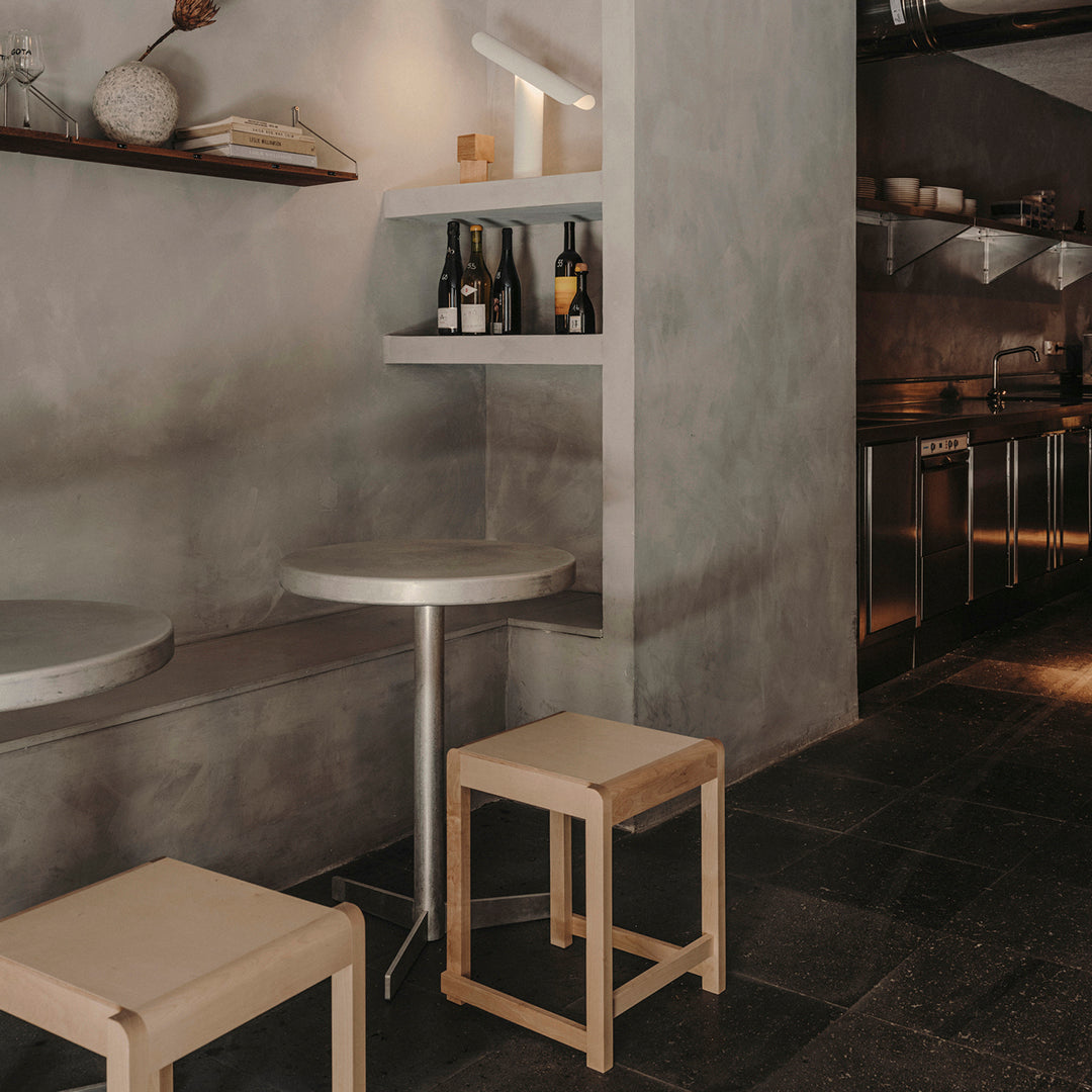 Minimalist interior featuring gray walls, sleek silver table, and light wooden stools against dark stone flooring.